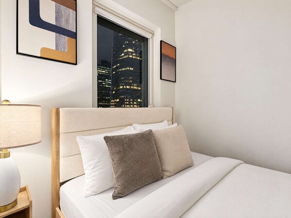 A hotel room with a bed, a nightstand, a lamp, and a window with a city view.