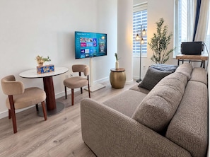 A modern living room with a flat-screen TV mounted on the wall, a round dining table with chairs, a sofa, and a potted plant.