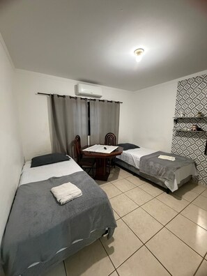 A room with two single beds, a table with chairs, an air conditioning unit, and patterned wallpaper.