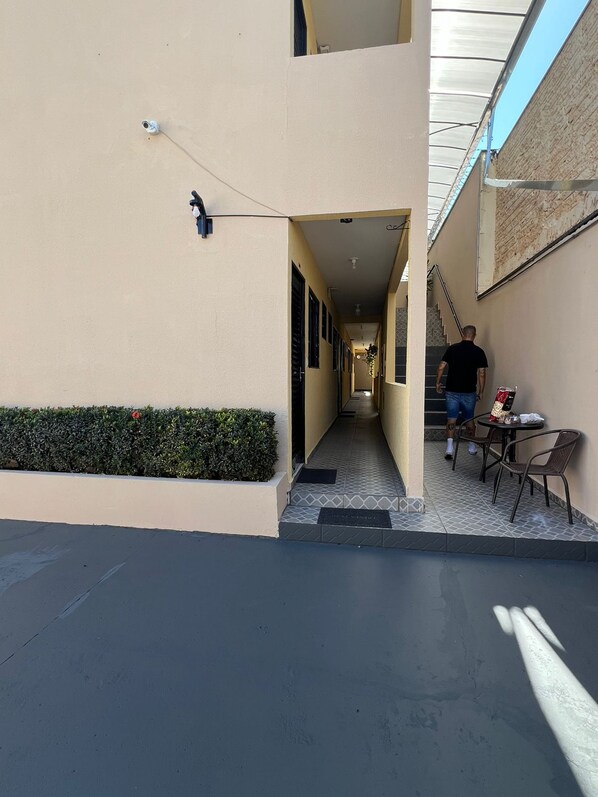 A narrow alleyway with a building on one side and a street on the other. There is a person walking down the alley, and a table with chairs outside the building.