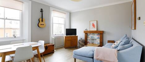 A living room with a blue sofa, a dining table with chairs, a guitar, a television, and a fireplace.