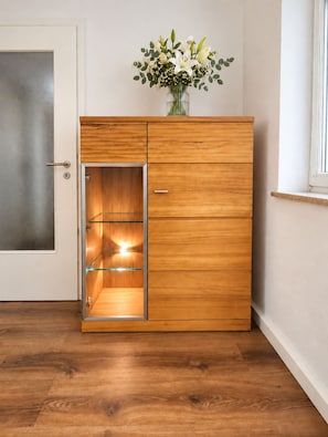 A wooden cabinet with a glass door, a vase with flowers on top, and a window to the right.
