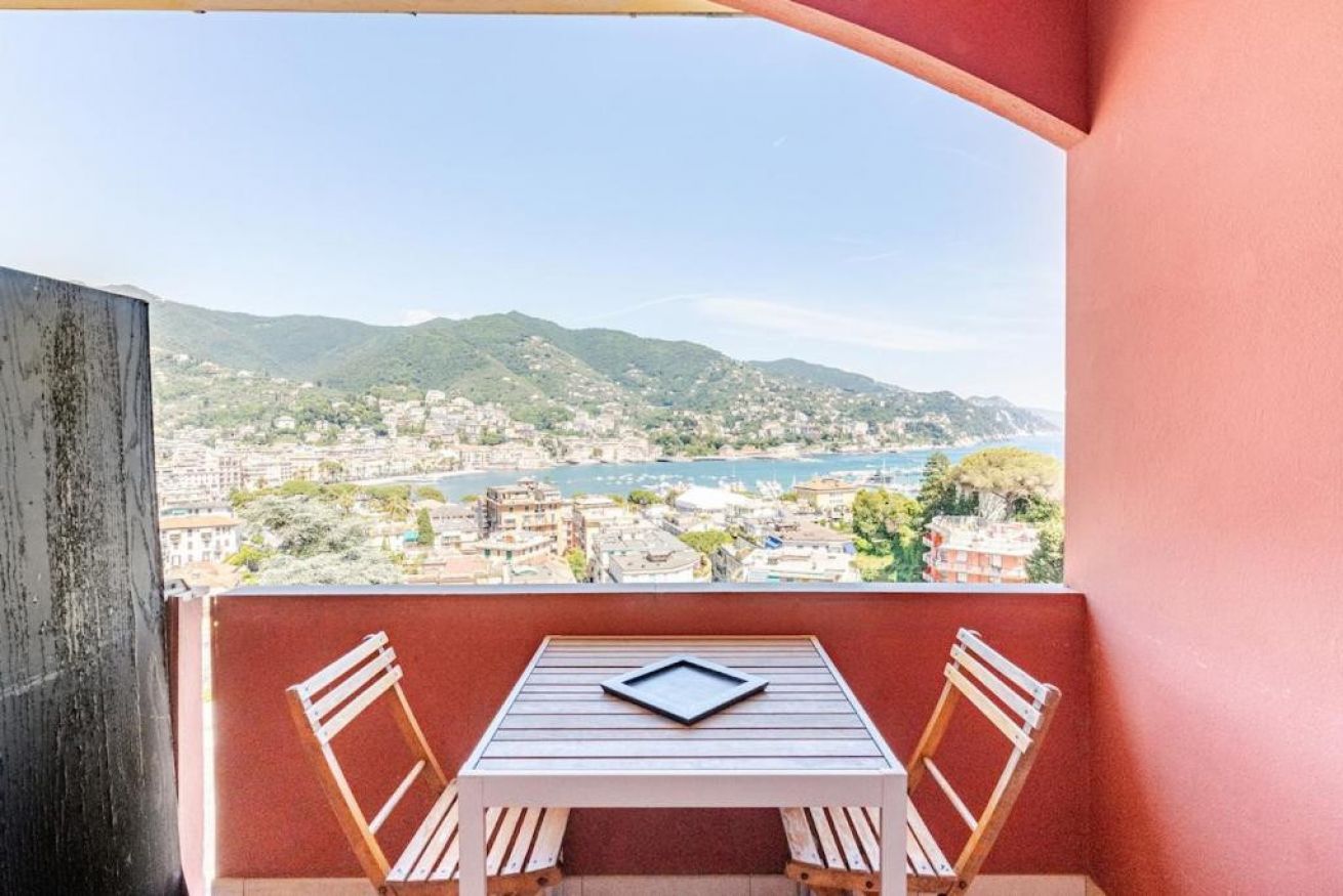 A balcony with a table and two chairs, overlooking a coastal cityscape with mountains in the distance.
