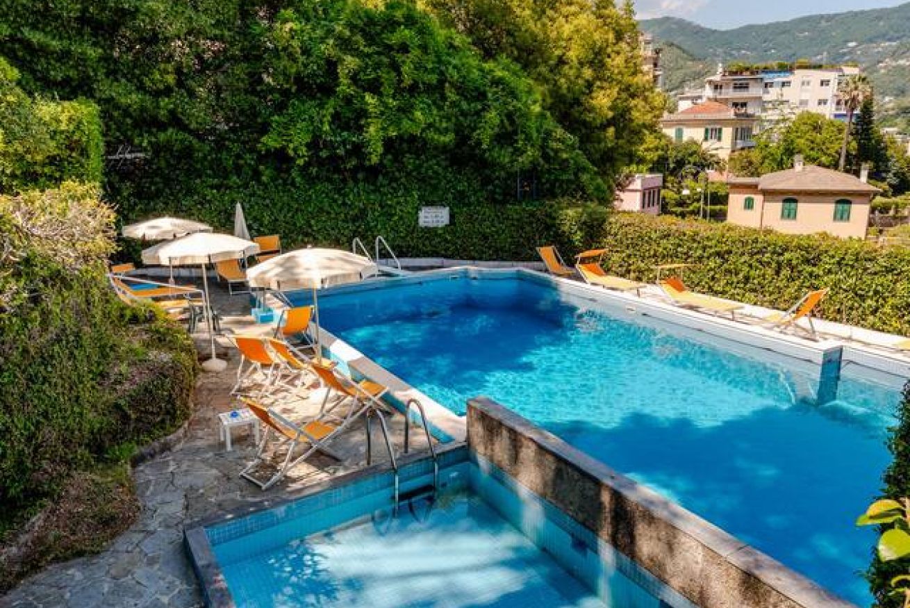 Una piscina rectangular con plataforma de piedra, rodeada de sillones de descanso naranjas y sombrillas, con un fondo de vegetación exuberante y edificios.