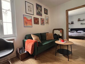 Una habitació amb un sofà verd, una taula rodona petita amb una teiera, i un mirall que reflecteix un llit amb roba de llit estampada.