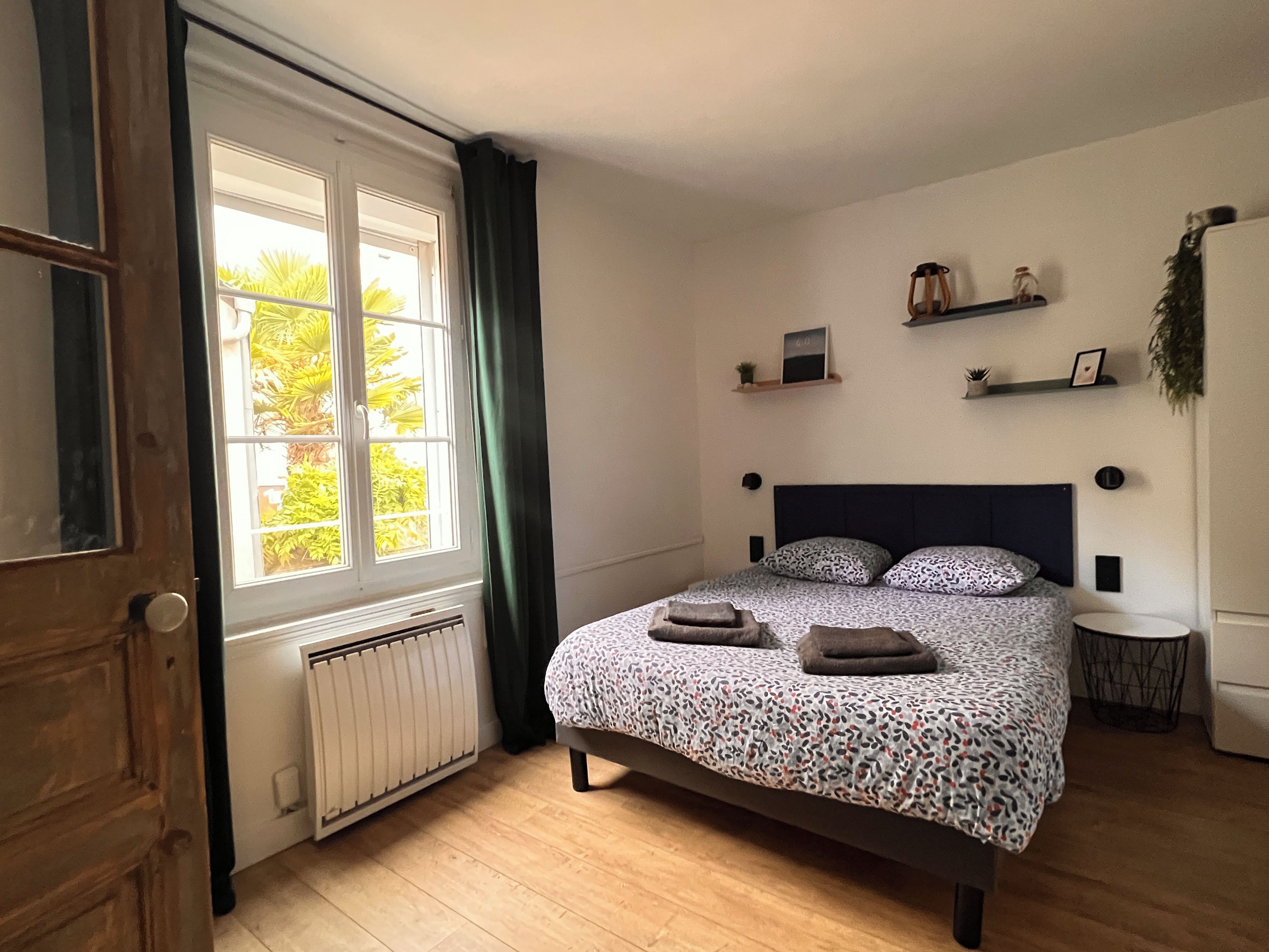 Een slaapkamer met een bed, een radiator, een raam met gordijnen en een plank met decoratieve objecten.