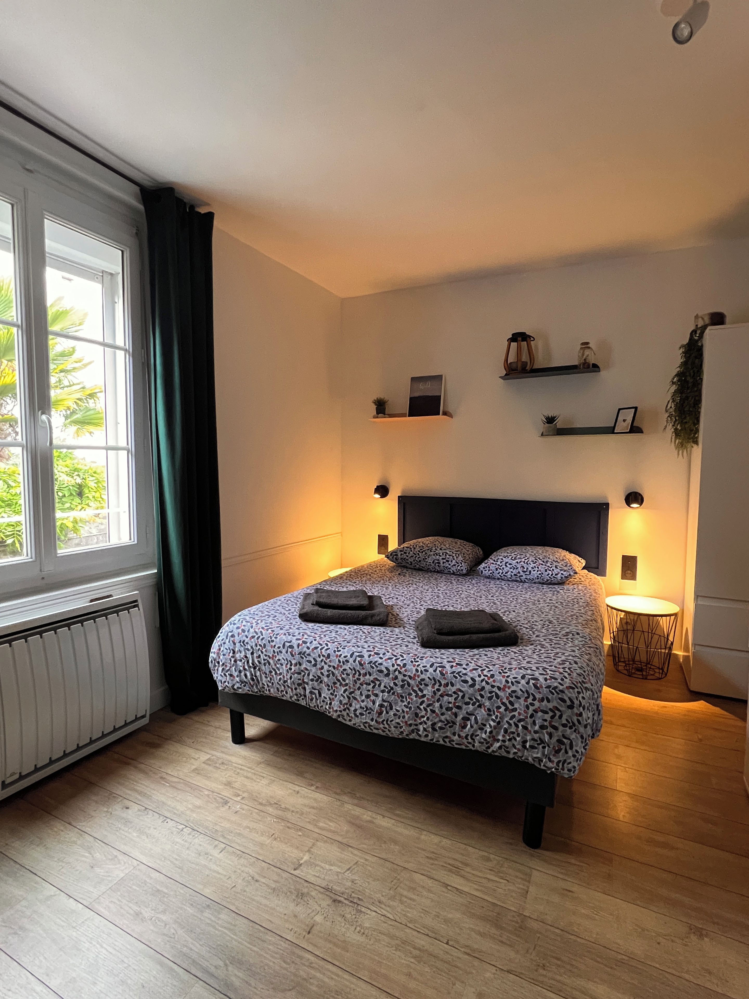 Une chambre à coucher avec un lit, des lampes de chevet, un radiateur, une fenêtre avec des rideaux et une petite table sur laquelle est posée une plante.
