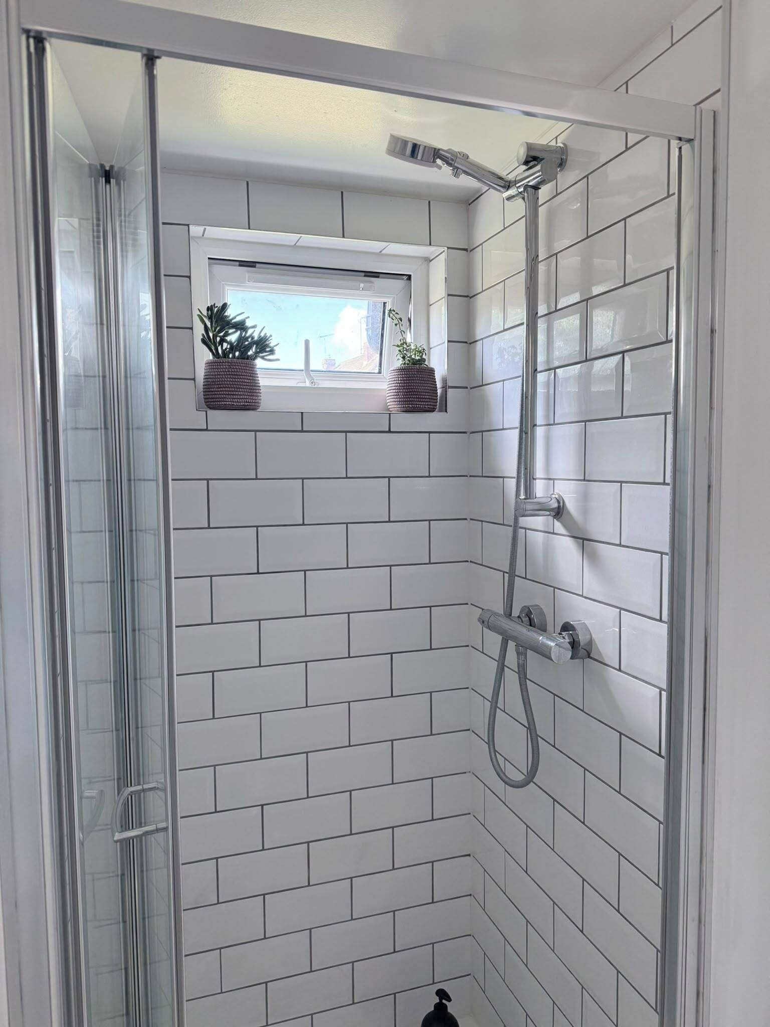 A shower with a glass door, white tiled walls, and a window with plants.