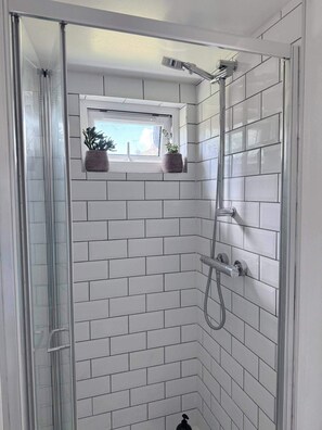 A shower with a glass door, white tiled walls, and a window with plants. - Cozy Cabin Retreat in Farnham (Farnham)