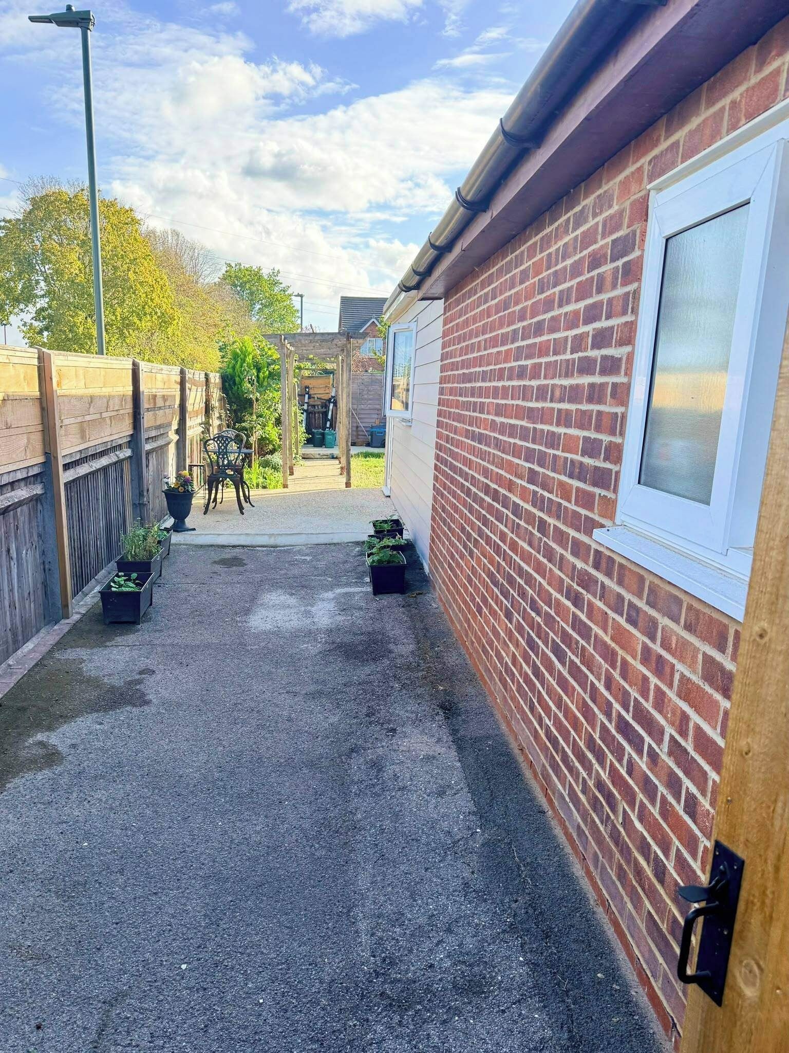 A narrow alleyway with a gravel path, wooden fence, and brick wall. There are potted plants and a wooden bench.