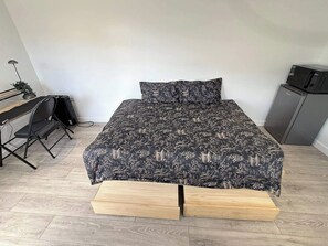 Uma cama com colcha estampada e almofadas, uma escrivaninha com cadeira, um pequeno armário e uma planta.
