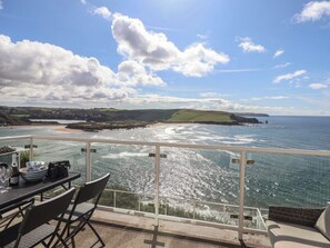 Ein Balkon mit Esstisch, der auf eine Küstenlandschaft mit Hügeln und dem Meer blickt.