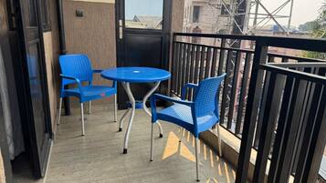 A balcony with a round table and two blue chairs, a black railing, and a view of buildings and a crane in the background.