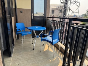 A balcony with a round table and two blue chairs, a black railing, and a view of buildings and a crane in the background.