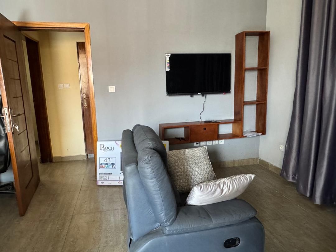A living room with a grey sofa, a mounted TV, a wooden shelf, and a door leading to another room.