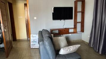 A living room with a grey sofa, a mounted TV, a wooden shelf, and a door leading to another room.