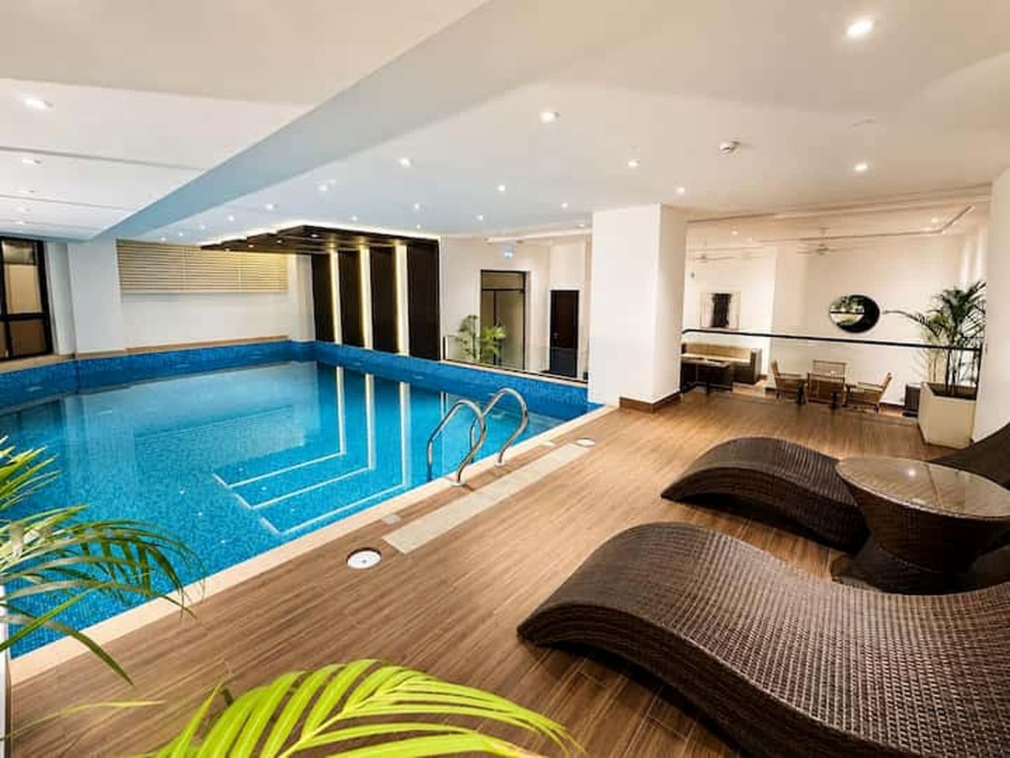 A modern indoor swimming pool with a wooden deck, lounge area, and a dining space.