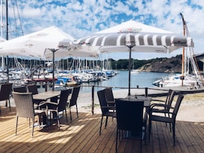 Un puerto deportivo con varias embarcaciones amarradas, mobiliario exterior con sombrillas y una terraza de madera.