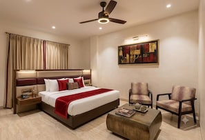 A hotel room with a bed, bedside tables, a coffee table, two armchairs, a ceiling fan, and a painting on the wall. - NewDelhi Hotel Citizen 1 (New Delhi)