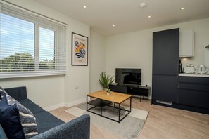 Ein modernes Wohnzimmer mit einer Couch, einem Couchtisch und einem Fernseher. Es gibt eine Küchenzeile mit einer Mikrowelle und einem Wasserkocher.