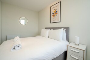 Habitación de hotel con una cama bien hecha, una mesita de noche con lámpara, un cuadro en la pared y un espejo redondo.
