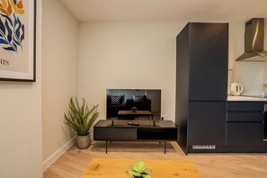 Ein modernes Wohnzimmer mit Flachbildfernseher, Couchtisch, Pflanze und einer Küchenzeile mit Herd und Schränken.
