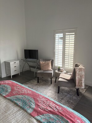 Ein Zimmer mit einem Bett, einem Schreibtisch mit Fernseher, zwei Stühlen, einem Fenster mit Jalousien und einem kleinen Tisch mit einer Pflanze.