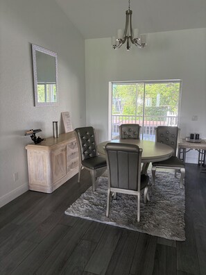 A dining area with a round table, four chairs, a chandelier, a mirror, and a window with a view of greenery. - Bonita Springs Paradise, Pool spa Canal home 5 min to city 20 min to beach & RSW (Bonita Springs)