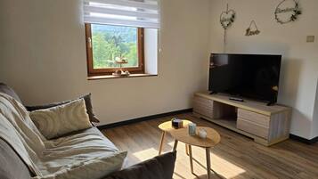 Una sala de estar con un sofá, un televisor sobre un mueble de madera, una mesita redonda con una taza y una ventana con persianas.