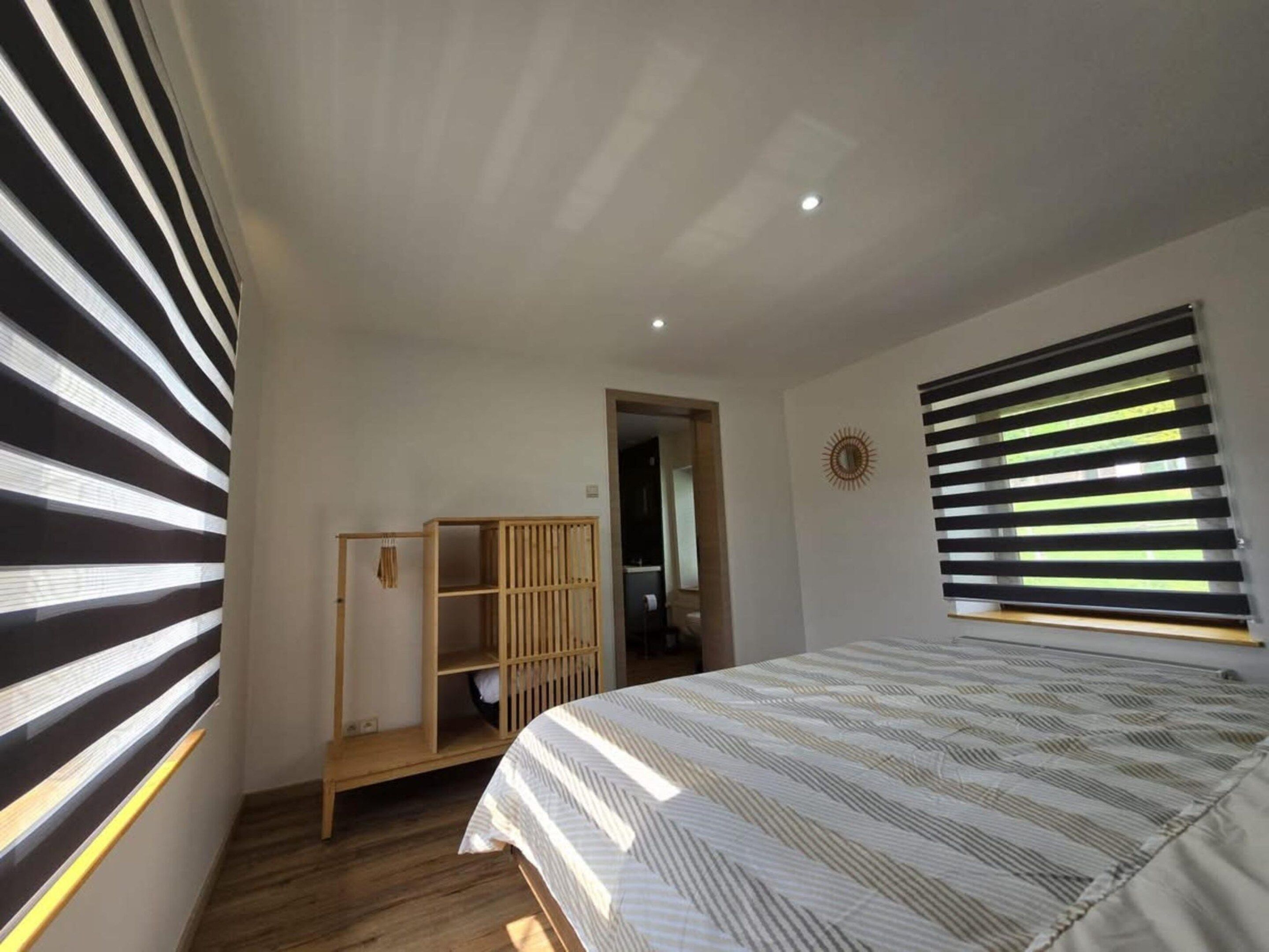 Ein Schlafzimmer mit einem Bett, einem Holzschrank, einem Fenster mit Jalousien und einer Tür zu einem anderen Raum.