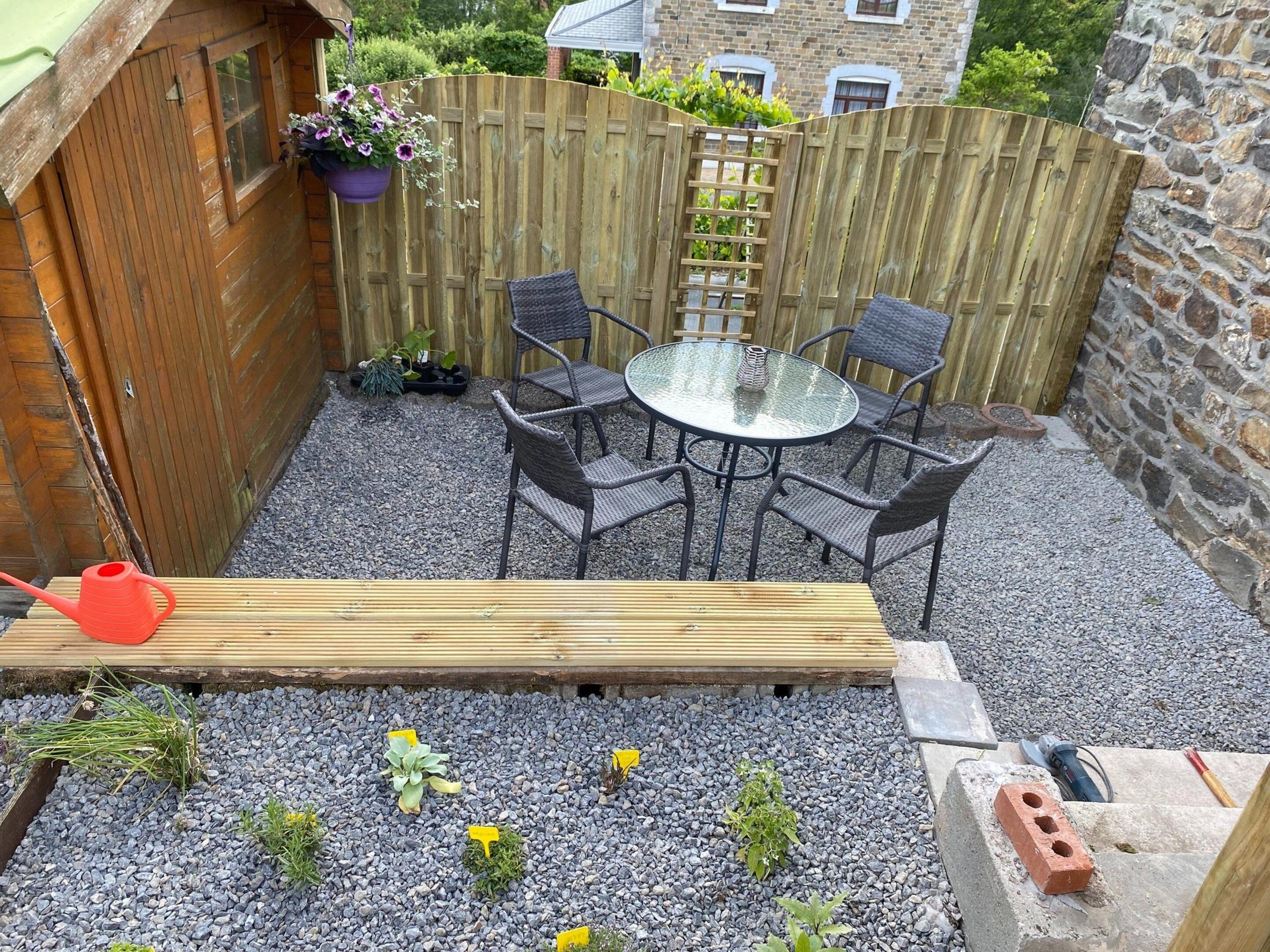 Una terraza de madera con una mesa redonda de vidrio y sillas, rodeada por una zona de grava con plantas.