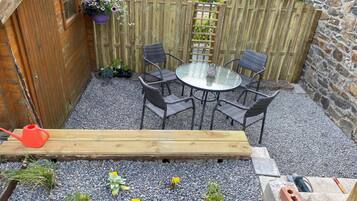 Una terraza de madera con una mesa redonda de vidrio y sillas, rodeada por una zona de grava con plantas.