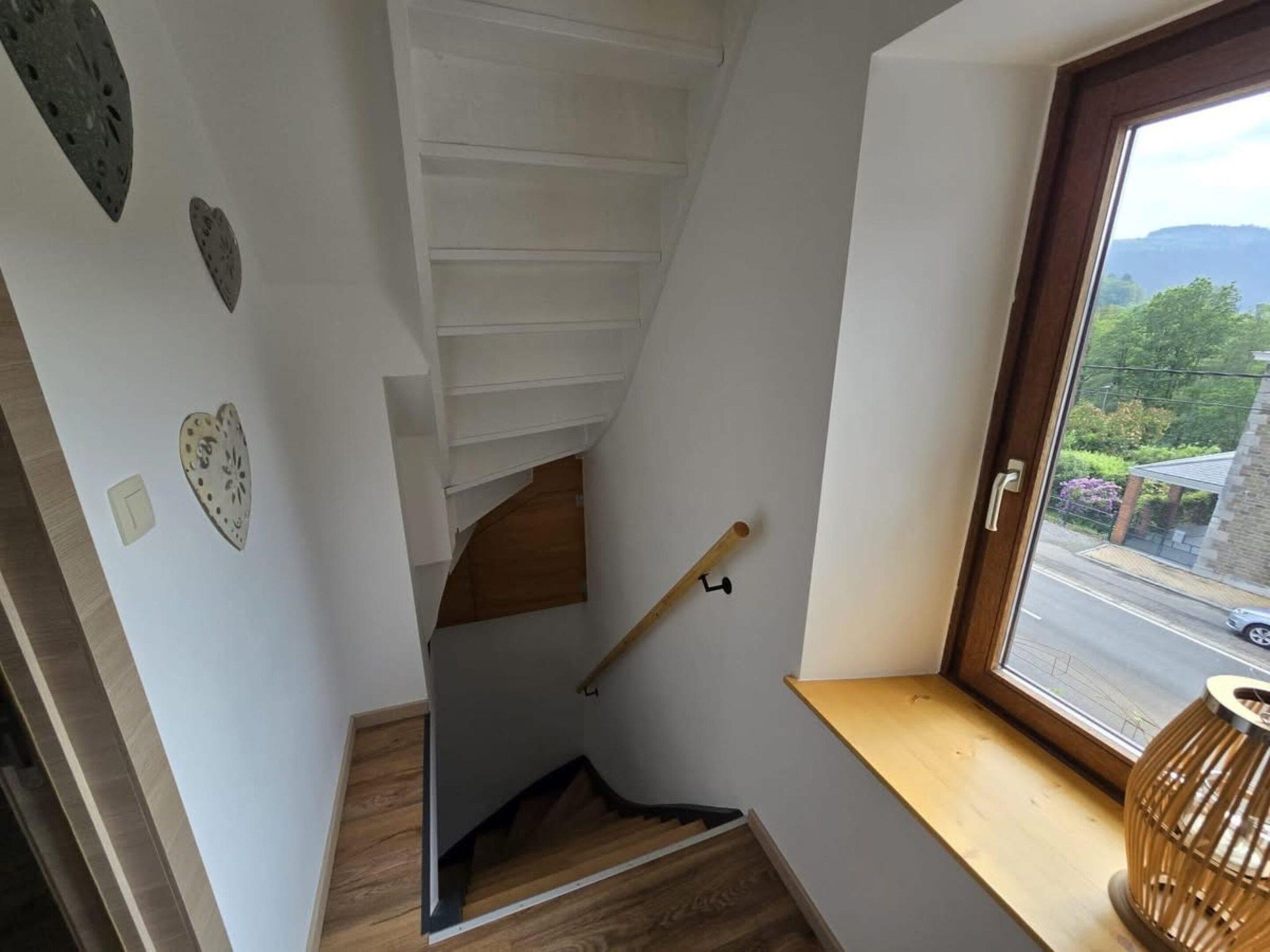 A staircase with a wooden handrail, white walls, and a window with a view of greenery and a building.