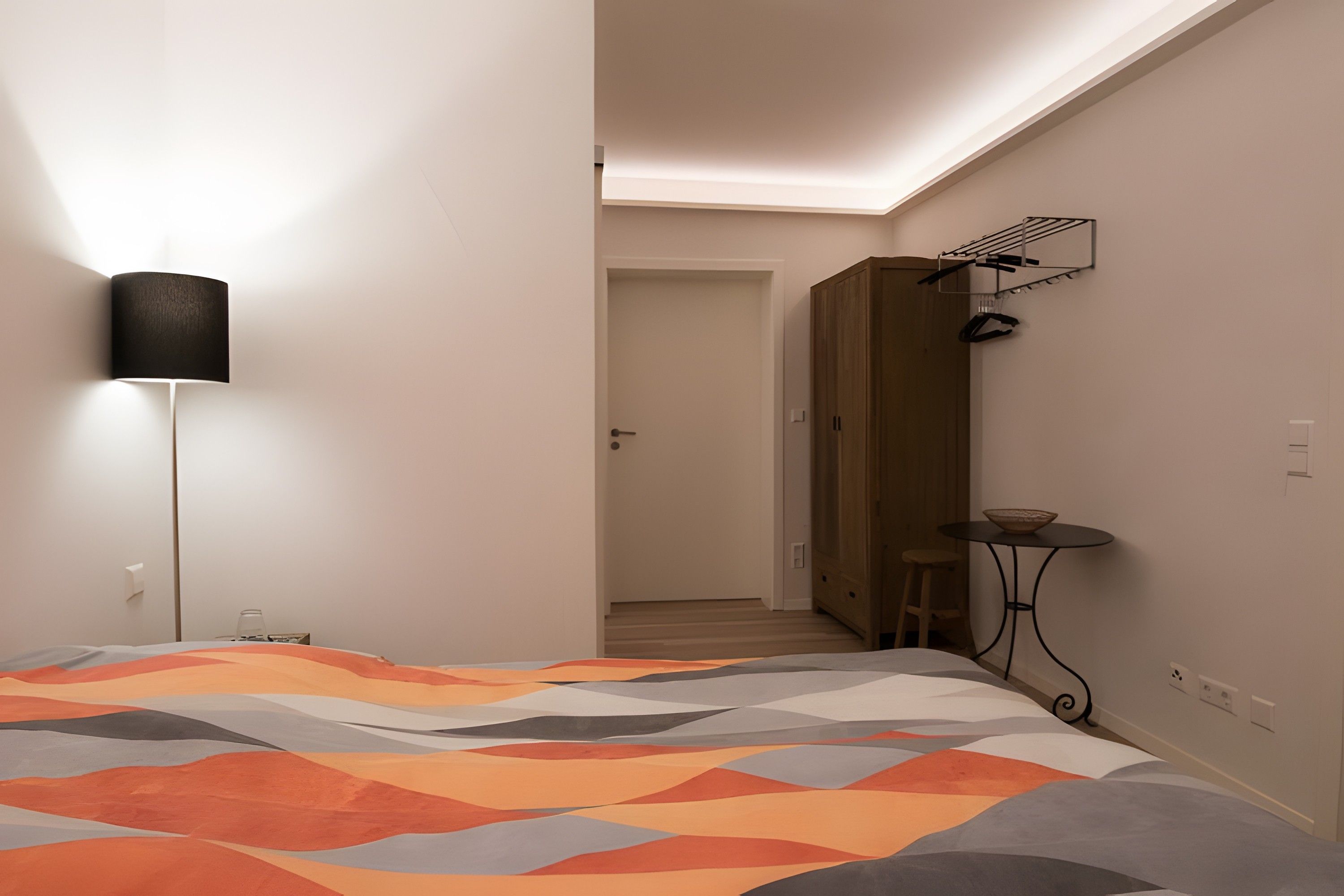 A bedroom with a bed, a bedside lamp, a wooden wardrobe, a small table with a bowl, and a chair.