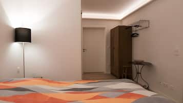 A bedroom with a bed, a bedside lamp, a wooden wardrobe, a small table with a bowl, and a chair.