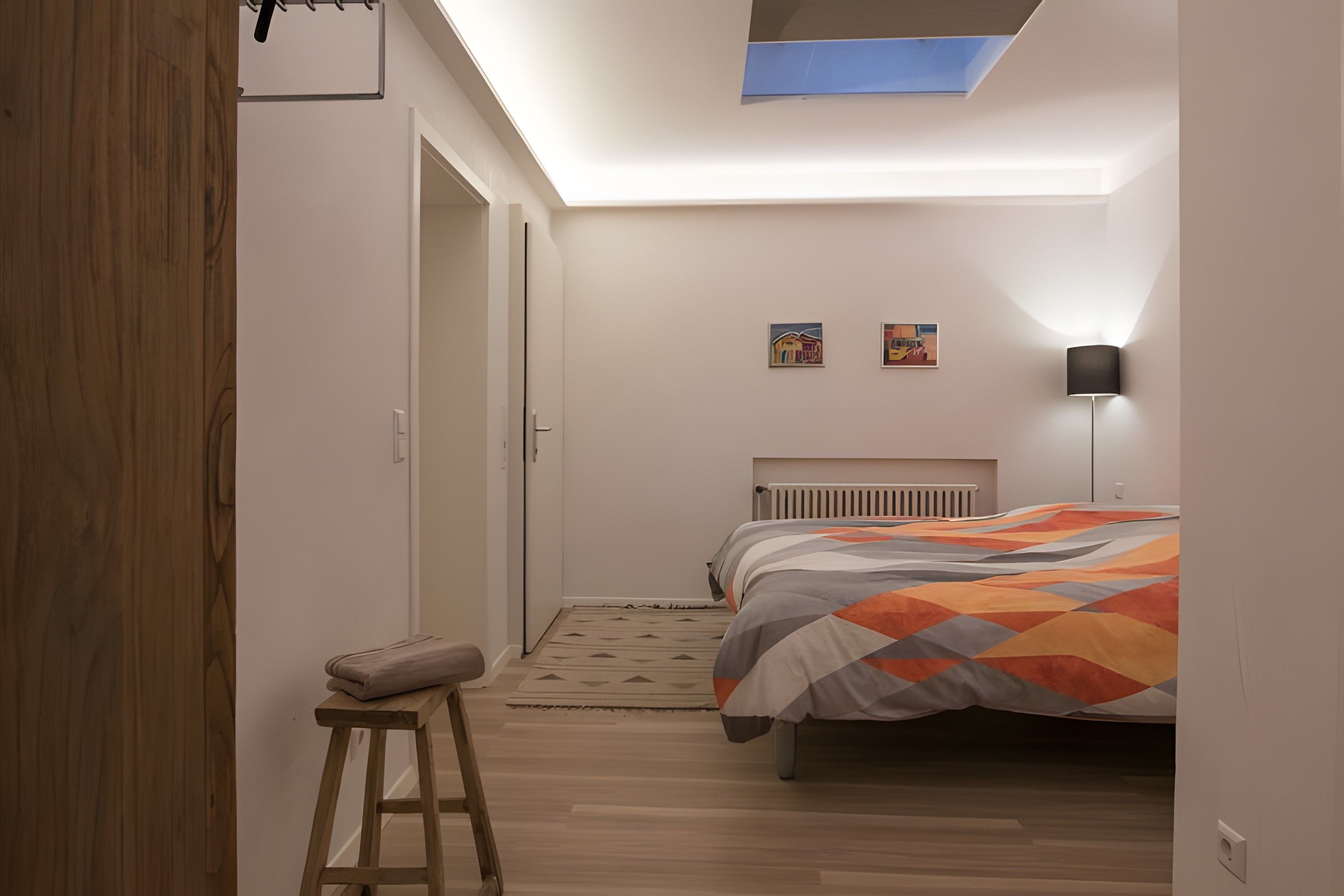 Un dormitorio con cama, mesita de noche, taburete, radiador y tragaluz.