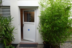 A white door with a blue accessibility symbol, a potted plant to the right, and a window with blinds partially drawn. - Apartment 'Großzügiges Ferienapartment' with Shared Garden – Central, Accessible (Kappelrodeck)