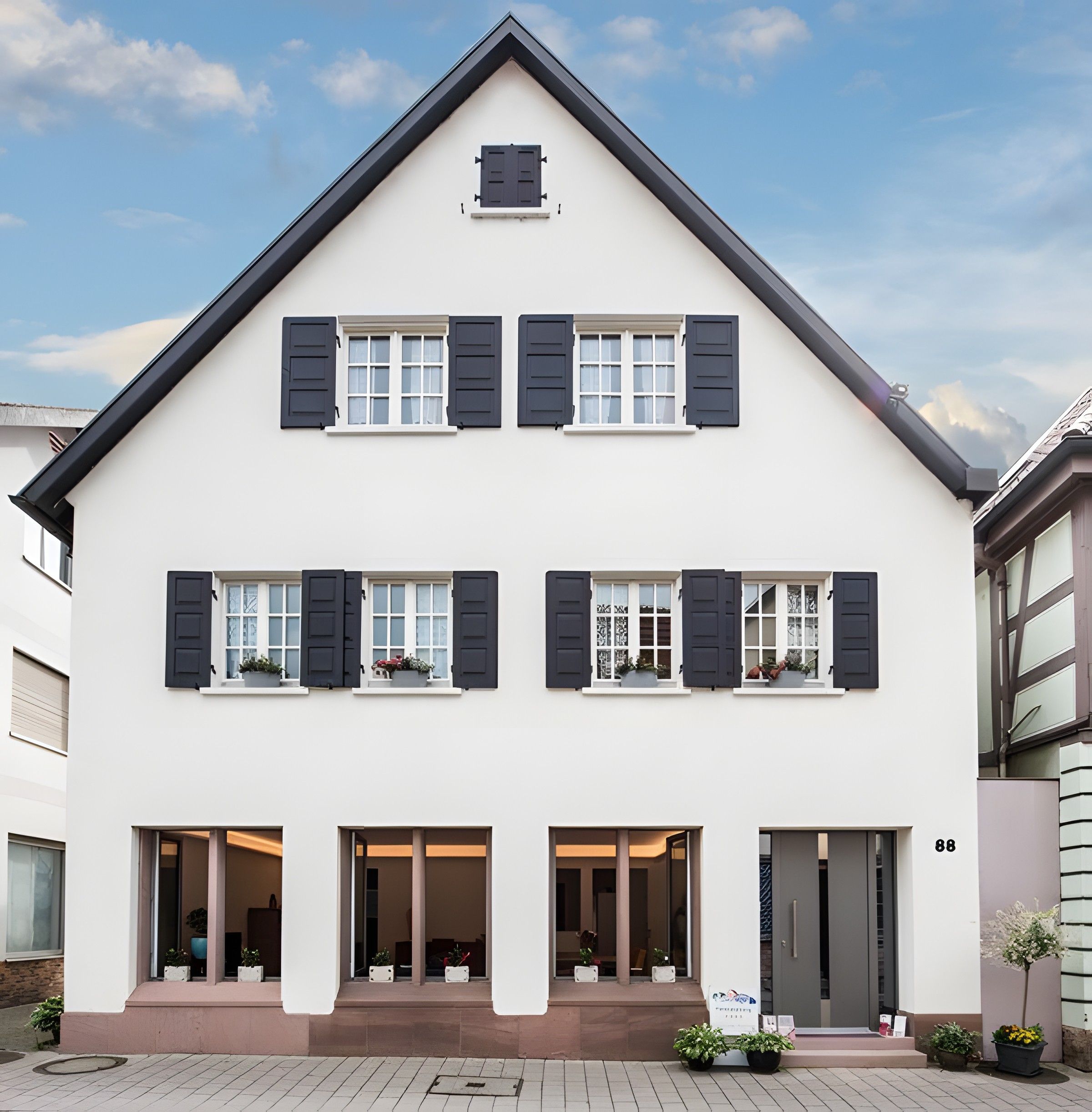 Ein zweistöckiges Gebäude mit einer symmetrischen Fassade, das einen zentralen Eingangsbereich mit einem überdachten Vorbau und zahlreiche Fenster mit Fensterläden aufweist.