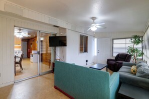 Ein Zimmer mit einer Schiebetür zu einer Küche, einem Deckenventilator, einem flachbildfernseher an der Wand und einer Sitzecke mit einer Couch und Sesseln.