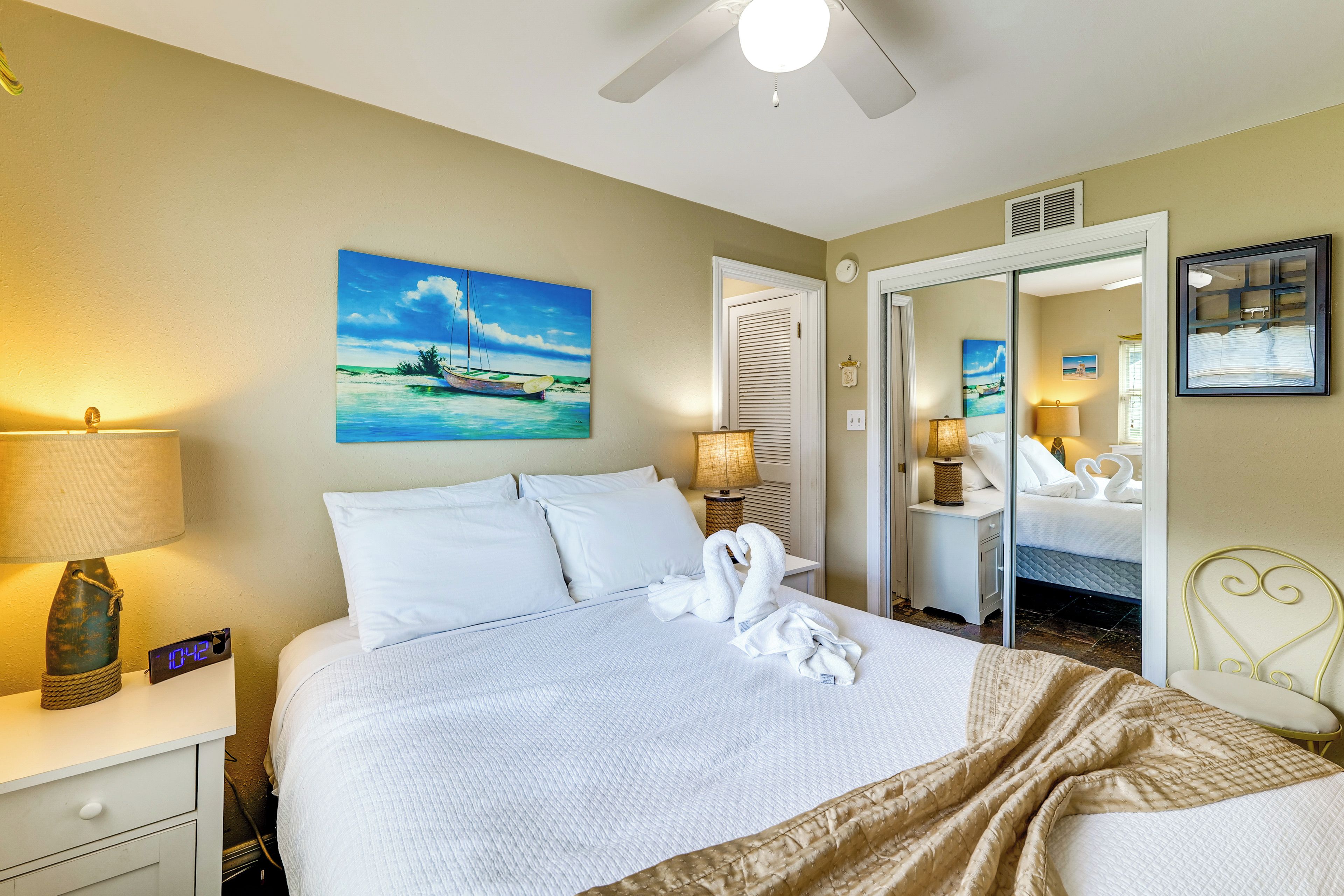 A hotel room with a bed, two bedside tables, a nightstand, a ceiling fan, and artwork on the walls.