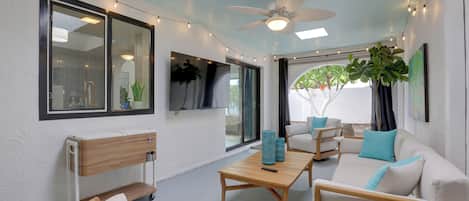 A modern living room with a ceiling fan, a TV mounted on the wall, and a large window providing a view of the outdoors.