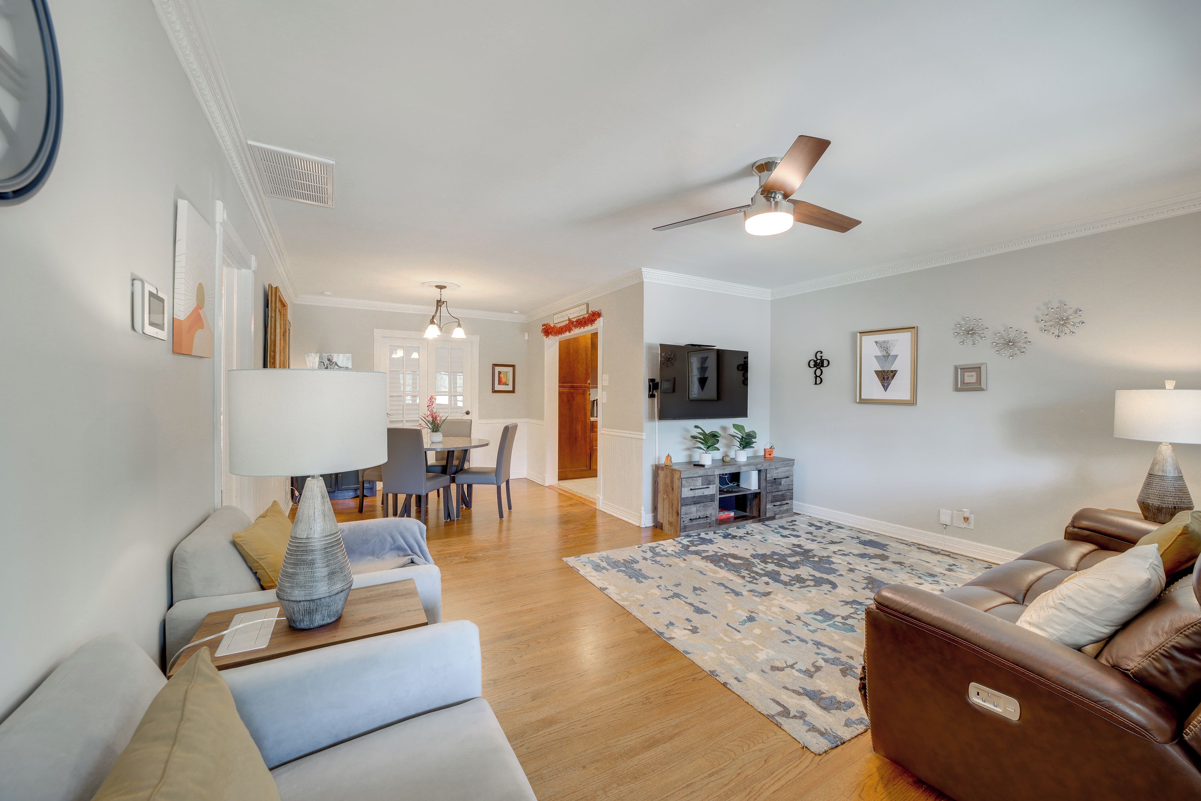 A modern living room with a sofa, a coffee table, and a leather armchair. There is a dining area with a table and chairs, and a kitchen area with a refrigerator and a TV mounted on the wall.