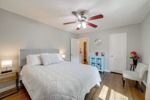 Ein Schlafzimmer mit einem großen Bett, einem Deckenventilator, einem Nachttisch mit Lampe, einem Schminktisch, einem Sessel und einer Tür.