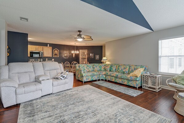 A living room with a grey sofa, a floral-patterned sofa, a dining area with wooden chairs, and a kitchen with a microwave. - Murrells Inlet Condo: Close to Beaches, Amenities (Murrells Inlet)
