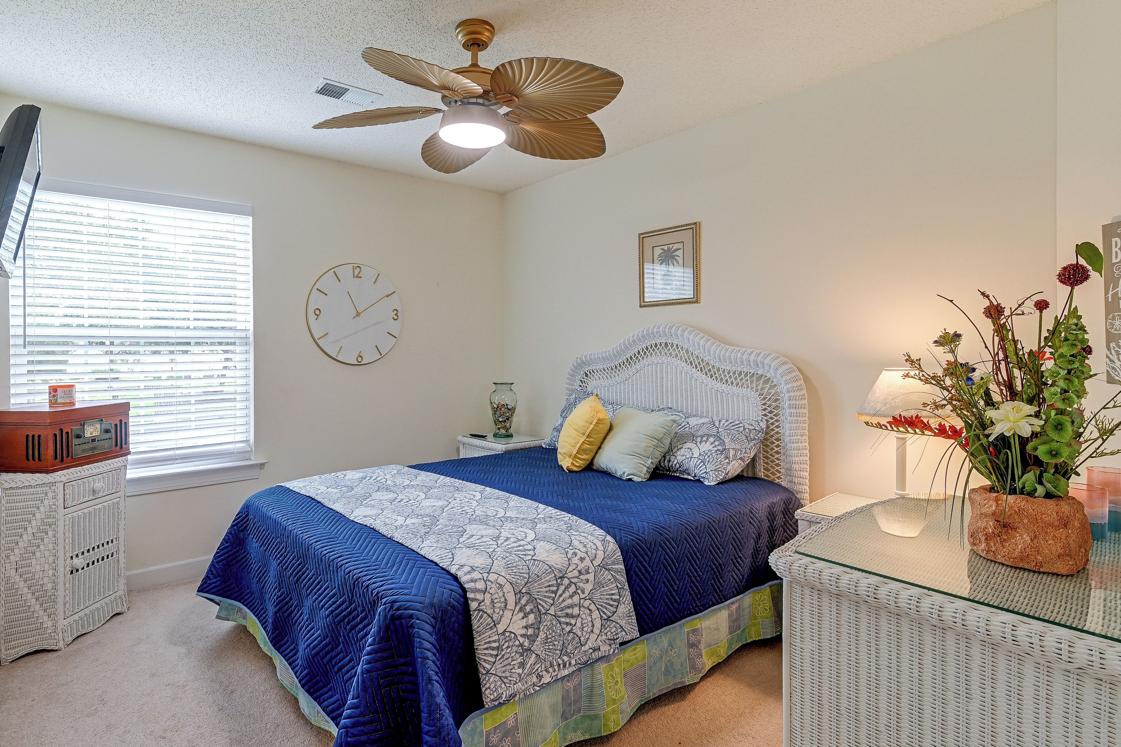 A bedroom with a bed, a ceiling fan, a clock, a window with blinds, a bedside table, and a nightstand.