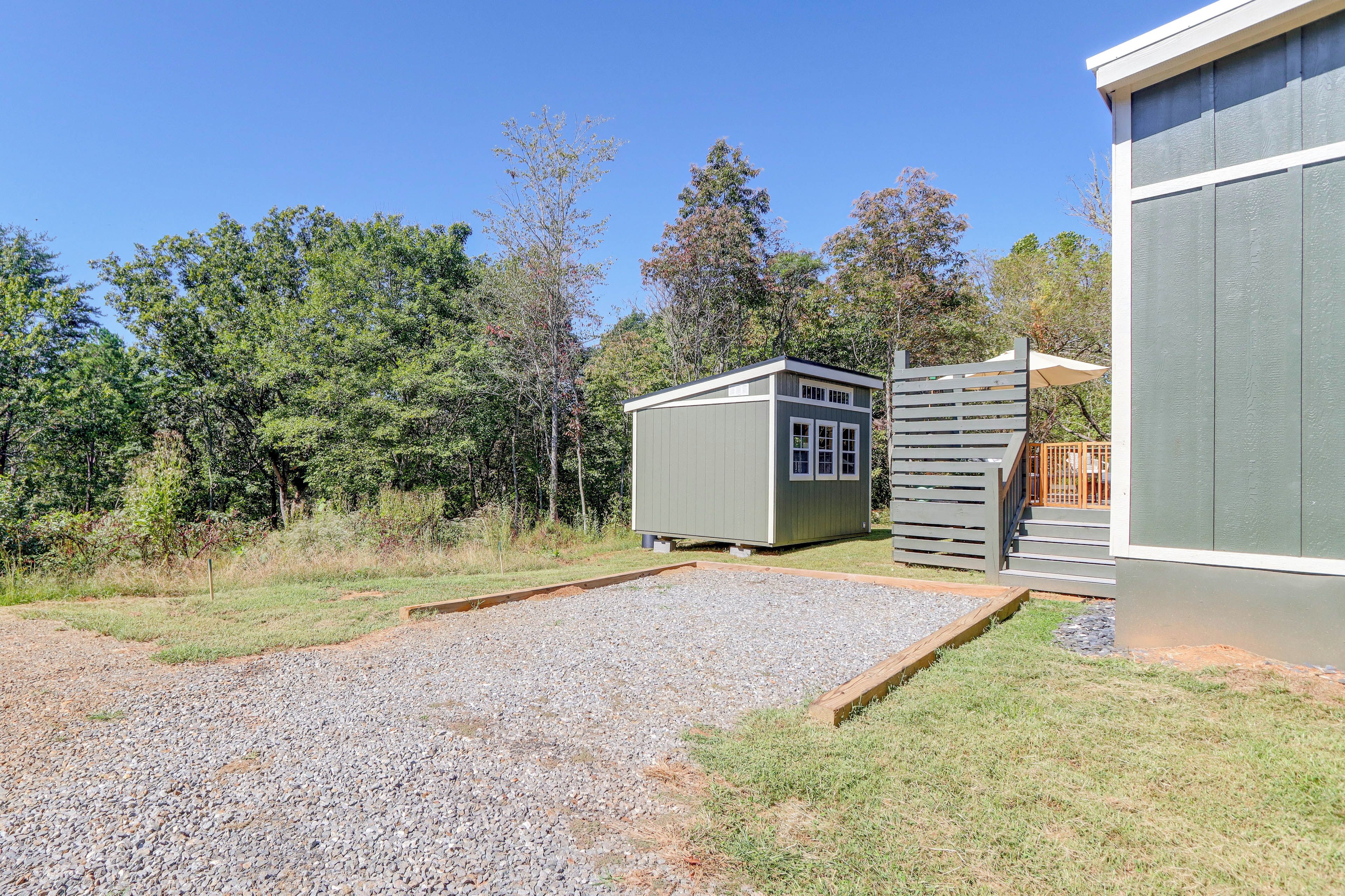 A gravel driveway leading to a small shed with a door and windows, surrounded by a wooden deck and a larger building with a sloped roof.