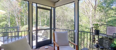 Una habitación soleada con ventanas amplias, una mesa de madera, dos sillas y una estantería metálica con plantas y una cita enmarcada.