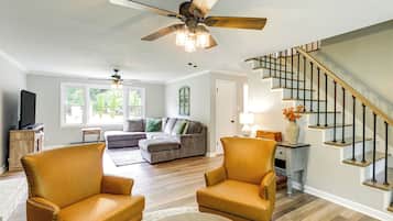 A living room with a sofa, two armchairs, a ceiling fan, a staircase, and a television.