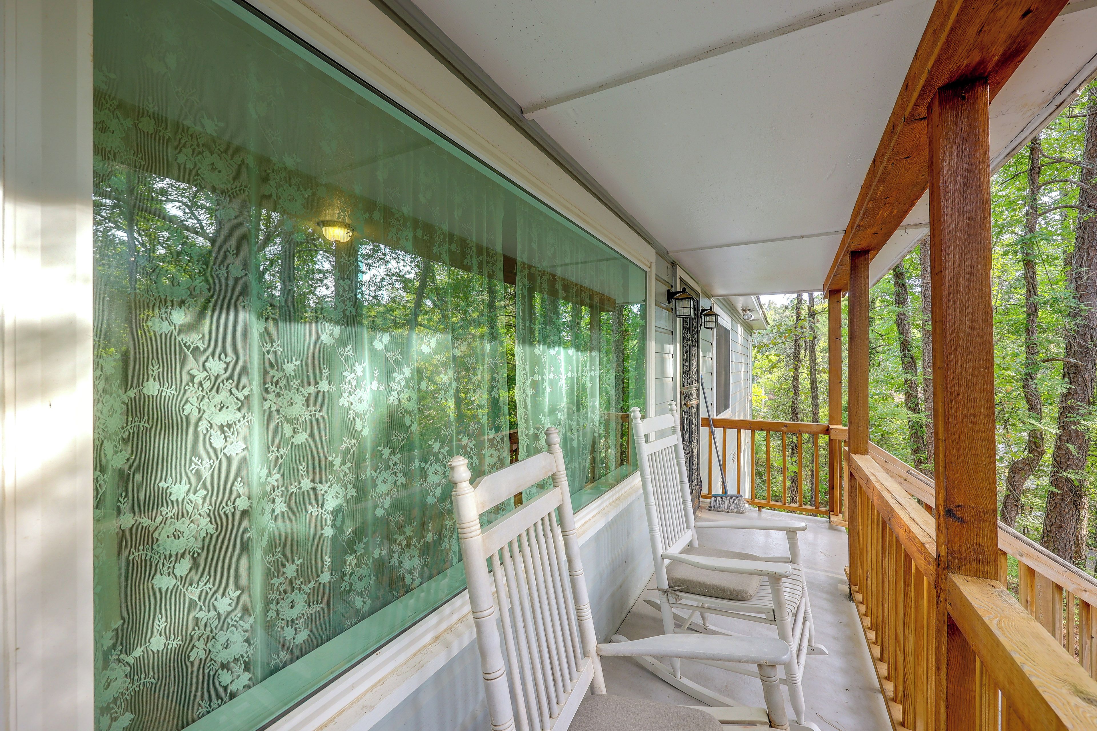 Une véranda couverte donnant sur une forêt, deux chaises blanches et une balustrade en bois.
