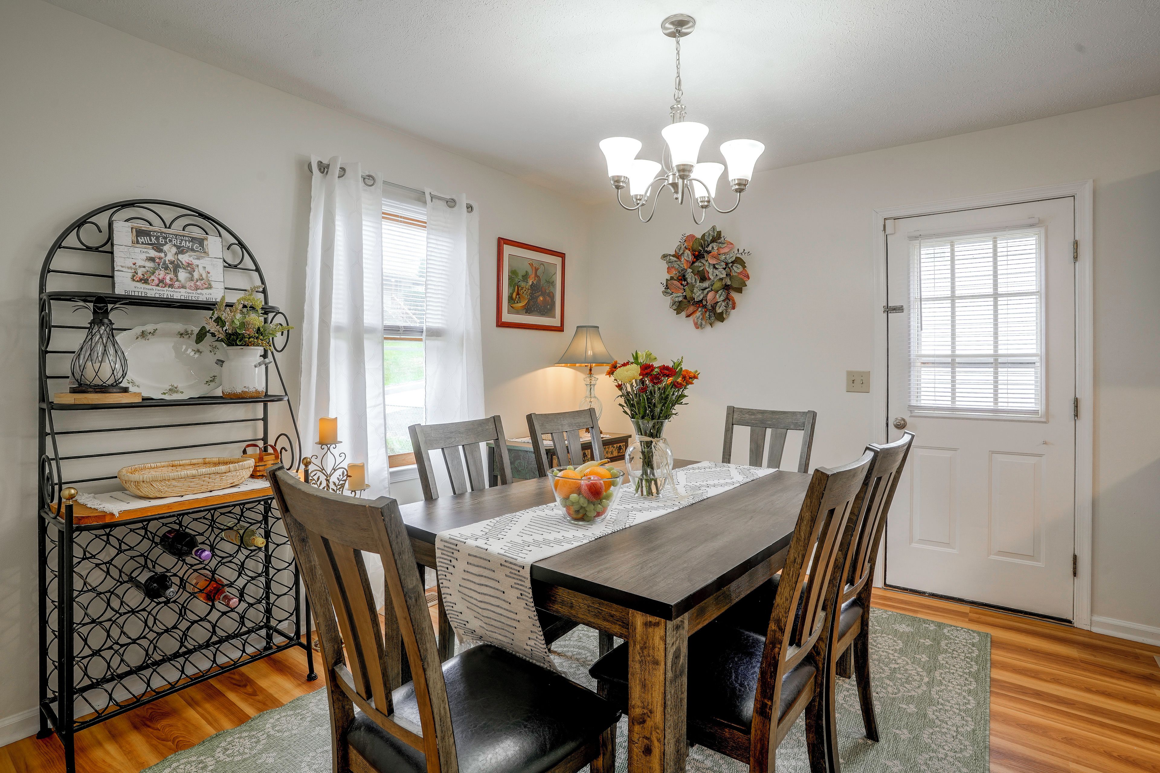 A dining room with a wooden table, chairs, a chandelier, a decorative wall piece, and a window with blinds.
